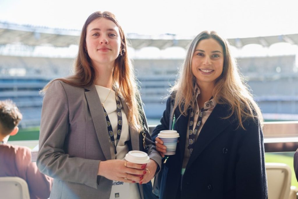 Two delegates with coffee cups smiling, the stadium seats and pitch behind them