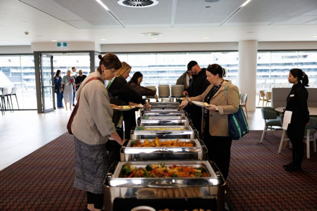 Delegates serve themselves lunch at the buffet in the stadium function room, windows behind